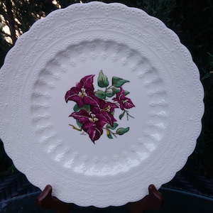 May include: White ceramic dinner plate with a raised, embossed border. The center of the plate features a hand-painted design of pink bougainvillea flowers.