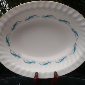 Op de afbeelding: Witte ovale serveerschaal met een gouden rand en een turquoise dolfijn- en bladerontwerp langs de rand. De schaal heeft een golvende, getextureerde rand. Een decoratief stuk voor het serveren van voedsel.