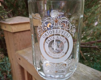 May include: A clear glass beer stein with a white and gold design. The design features the text "Oktoberfest Paulaner Munchen".