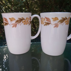 May include: Two white ceramic mugs with a gold rim and a brown and yellow floral design. The mugs are on a glass table.