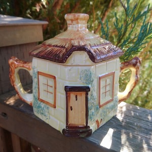 Pode incluir: Uma chaleira de cerâmica em forma de casa de campo. A chaleira tem uma tampa com telhado de colmo, um corpo creme com detalhes em tijolo e detalhes castanhos. Possui uma porta, janelas e alças. É um item decorativo.