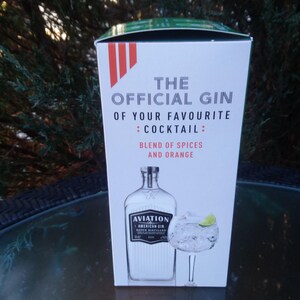 May include: A white box with the text "THE OFFICIAL GIN OF YOUR FAVOURITE : COCKTAIL : BLEND OF SPICES AND ORANGE." The box features an illustration of a gin bottle and a cocktail glass with a lime wedge. The gin bottle is labeled "AVIATION AMERICAN GIN."