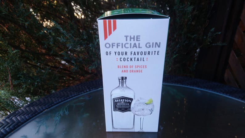May include: A white box with the text "THE OFFICIAL GIN OF YOUR FAVOURITE : COCKTAIL : BLEND OF SPICES AND ORANGE." The box features an illustration of a gin bottle and a cocktail glass with a lime wedge. The gin bottle is labeled "AVIATION AMERICAN GIN."