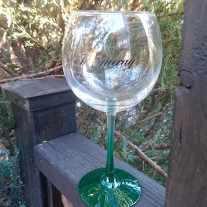 Op de afbeelding: Een helder wijnglas met een groene steel en voet, rustend op een houten leuning. Het woord "Tanqueray" is in het zwart op het glas gedrukt. De achtergrond toont groene bladeren en boomtakken.