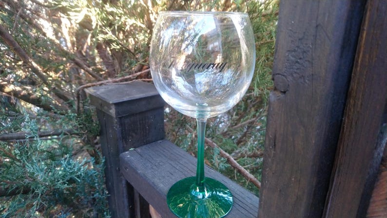 May include: A clear wine glass with a green stem and base, resting on a wooden railing. The word "Tanqueray" is printed on the glass in black. The background features green foliage and tree branches.