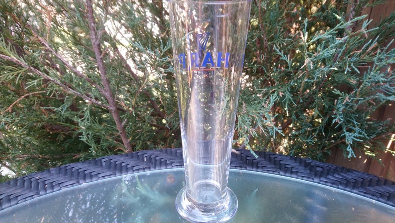 Może przedstawiać: Szklany kufel do piwa z przezroczystego szkła z niebiesko-złotym logo z napisem "Pilsner Urquell" z przodu.