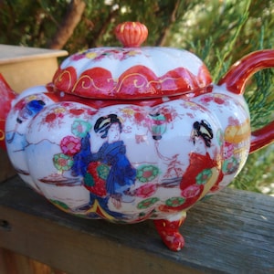 Puede incluir: Una tetera blanca con tapa y asa rojas, decorada con diseños florales y geishas en rojo, azul y dorado. La tetera está sobre una superficie de madera.