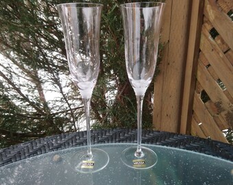 Può includere: Due flute da champagne in vetro trasparente con un'etichetta dorata che recita "Mikasa" sul fondo di ogni bicchiere. Le flute sono appoggiate su un tavolo in vimini nero.
