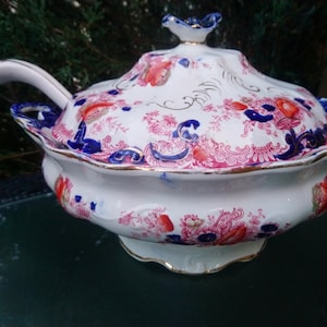 May include: A white porcelain soup tureen with a lid and a ladle. The tureen is decorated with a floral pattern in shades of pink, blue, and gold.