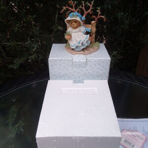 Pode incluir: Uma figurinha de porcelana branca de um urso vestindo um vestido azul e um chapéu branco, segurando um ramo de cenouras. A figurinha está em pé sobre uma base com uma pequena cerca e uma árvore. A base é decorada com um padrão floral azul e branco.