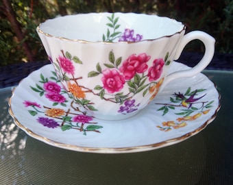May include: A white teacup and saucer with a floral pattern. The cup and saucer have a gold trim and feature pink, purple, yellow, and orange flowers with green leaves. A small bird is painted on the saucer.