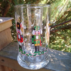 May include: A clear glass beer stein with a Christmas theme. The stein features three nutcracker figures with red, green, and gold accents. The figures are surrounded by holly and snowflakes.
