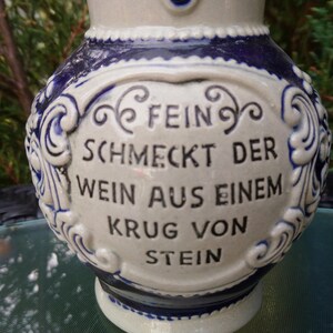 Puede incluir: Una jarra de cer&aacute;mica blanca con dise&ntilde;os florales azules y blancos. La jarra tiene una cita en alem&aacute;n que dice "Fein schmeckt der Wein aus einem Krug von Stein".