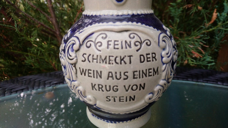 Puede incluir: Una jarra de cer&aacute;mica blanca con dise&ntilde;os florales azules y blancos. La jarra tiene una cita en alem&aacute;n que dice "Fein schmeckt der Wein aus einem Krug von Stein".