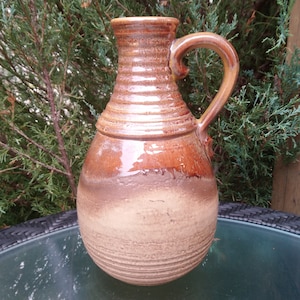 May include: A brown ceramic pitcher with a wide, round body and a handle. The pitcher has a glazed finish and a textured surface.
