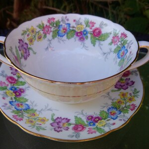 May include: A white porcelain soup bowl with a floral pattern in shades of pink, yellow, and blue. The bowl has a gold rim and is sitting on a matching saucer.