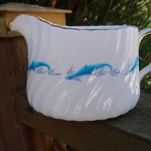 May include: White ceramic creamer with a gold rim and blue dolphin design. The creamer is sitting on a wooden surface.