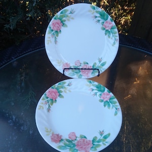 May include: Two white ceramic plates with a floral rose design. The plates have a white background with a border of pink roses and green leaves. The plates are displayed on a glass table.