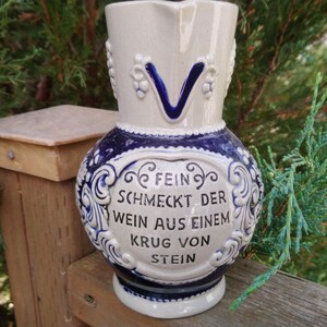 Puede incluir: Una jarra de cer&aacute;mica blanca y azul con asa y un dise&ntilde;o floral azul y blanco. La jarra tiene una cita en alem&aacute;n que dice "Fein schmeckt der Wein aus einem Krug von Stein." que se traduce como "El vino de una jarra de piedra sabe bien."