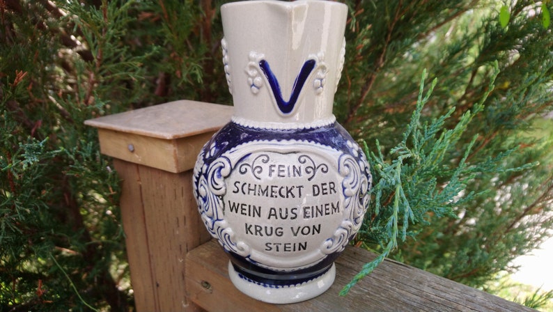 Puede incluir: Una jarra de cer&aacute;mica blanca y azul con asa y un dise&ntilde;o floral azul y blanco. La jarra tiene una cita en alem&aacute;n que dice "Fein schmeckt der Wein aus einem Krug von Stein." que se traduce como "El vino de una jarra de piedra sabe bien."