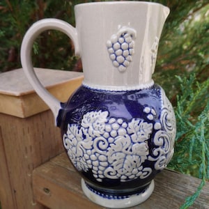 Puede incluir: Una jarra de cer&aacute;mica azul y blanca con asa y dise&ntilde;o de uvas. La jarra est&aacute; decorada con un dise&ntilde;o de uvas blancas sobre fondo azul.