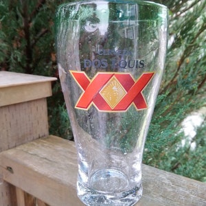 May include: A clear glass beer mug with the logo "Cerveza Dos Equis XX" printed on the front. The logo is red, gold, and white.