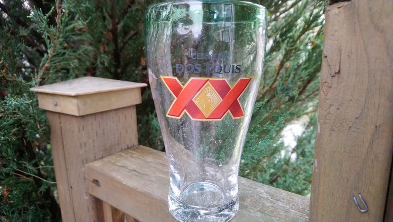 May include: A clear glass beer mug with the logo "Cerveza Dos Equis XX" printed on the front. The logo is red, gold, and white.