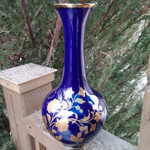 May include: A cobalt blue ceramic vase with a gold rim and intricate gold floral designs. The vase has a round body and a long, slender neck.