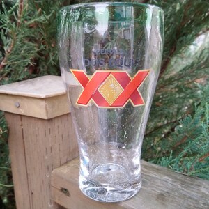 May include: A clear glass beer mug with a red and gold Dos Equis logo. The mug is sitting on a wooden surface.