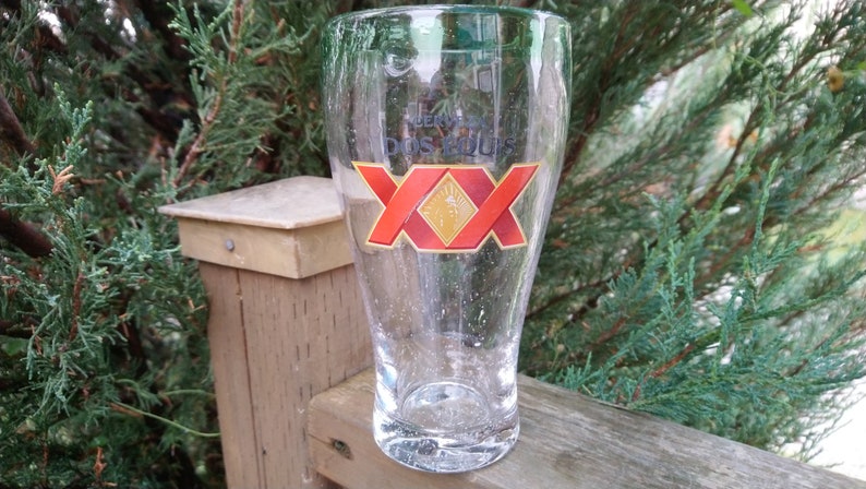 May include: A clear glass beer mug with a red and gold Dos Equis logo. The mug is sitting on a wooden surface.