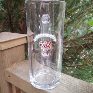 Può includere: Un boccale di birra in vetro trasparente con manico. Il boccale ha un'etichetta blu e dorata con il testo "Himmel der Bayern" e "Hacker-Pschorr Munchen".