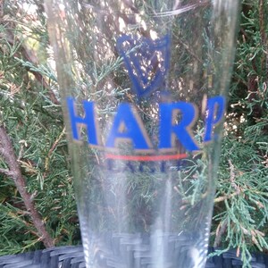 Może przedstawiać: Szklany kufel do piwa o pojemności pinta, z niebiesko-złotym logo Harp Lager. Logo przedstawia harfę i napis "Harp Lager".