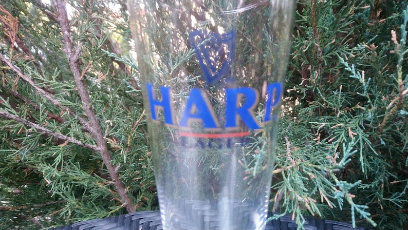 Może przedstawiać: Szklany kufel do piwa o pojemności pinta, z niebiesko-złotym logo Harp Lager. Logo przedstawia harfę i napis "Harp Lager".