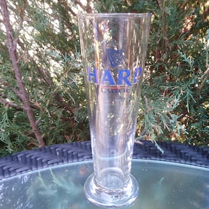 Może przedstawiać: Szklany kufel do piwa z przezroczystego szkła z niebiesko-białym logo Harp Lager. Kufel stoi na czarnym wiklinowym stole.