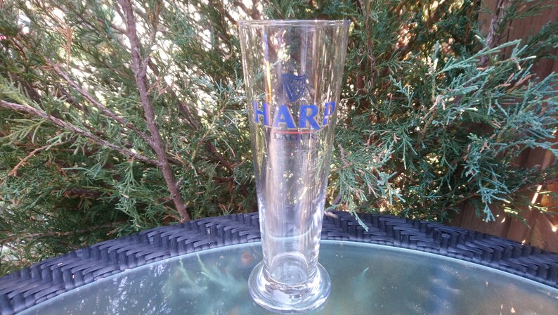 Może przedstawiać: Szklany kufel do piwa z przezroczystego szkła z niebiesko-białym logo Harp Lager. Kufel stoi na czarnym wiklinowym stole.