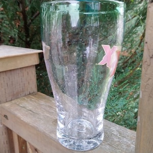 May include: A clear glass pint glass with a pink and white logo on the side. The glass is sitting on a wooden surface.