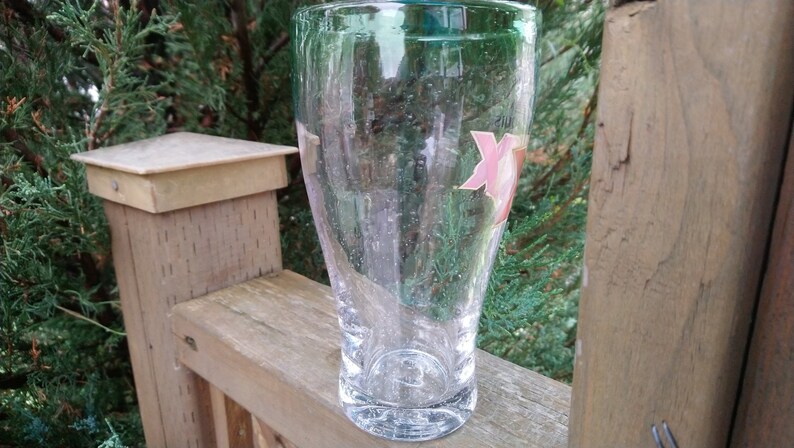 May include: A clear glass pint glass with a pink and white logo on the side. The glass is sitting on a wooden surface.
