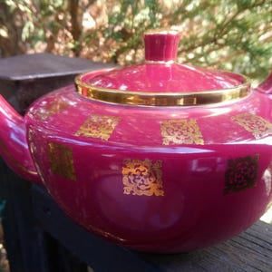 May include: A deep crimson red teapot with a gold trim and gold patterned design. The teapot has a lid and a handle.