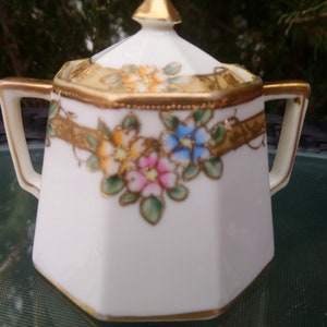 May include: White porcelain sugar bowl with a lid, featuring a floral design with gold accents. The bowl has two handles and a hexagonal shape.