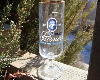 Op de afbeelding: Een helder glazen bierglas met een gouden rand. Het glas heeft een blauw en wit logo met de tekst "J.B. Degroof Pilsner" en de jaren "1858" en "1958".