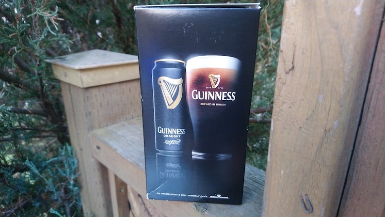 May include: A black box with a can and a glass of Guinness beer. The can says "Guinness Draught" and the glass says "Guinness Brewed in Dublin".