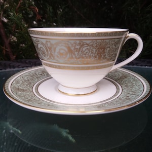 May include: A white teacup and saucer with a green and gold floral pattern. The cup has a handle and the saucer has a scalloped edge.