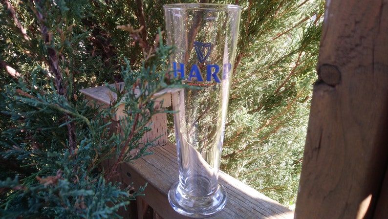 Może przedstawiać: Szklany kufel do piwa z przezroczystego szkła z niebiesko-białym logo Harp. Kufel stoi na drewnianej powierzchni.