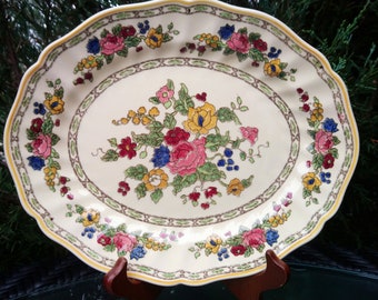 May include: A white oval serving platter with a scalloped edge. The platter is decorated with a floral pattern in shades of pink, yellow, blue, and green. The floral design features a central bouquet of roses and other flowers.