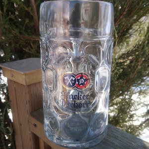 May include: A clear glass beer stein with a textured surface. The stein has a logo with the text "Hacker-Pschorr" and two symbols, one with a crossed axe and the other with a star.
