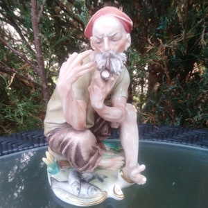 May include: A porcelain figurine of a man with a red hat, sitting on a base with fish. He is wearing a brown shirt and pants and has a white beard. He is holding a pipe in his hand.