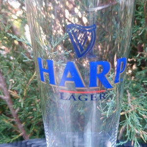 Może przedstawiać: Szklany kufel do piwa z przezroczystego szkła z nadrukowanym z przodu logo Harp Lager. Logo jest niebieskie i złote i przedstawia harfę.