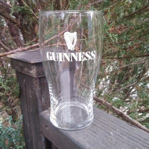 Puede incluir: Un vaso de pinta de vidrio transparente con el logotipo de Guinness y un dise&ntilde;o de arpa impreso en la parte delantera. El vaso est&aacute; sobre una superficie de madera.
