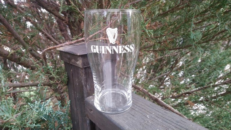 Puede incluir: Un vaso de pinta de vidrio transparente con el logotipo de Guinness y un dise&ntilde;o de arpa impreso en la parte delantera. El vaso est&aacute; sobre una superficie de madera.
