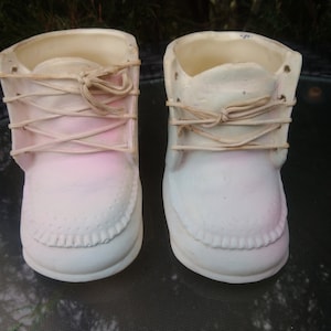 Può includere: Un paio di scarpine da bebé in ceramica bianca con dettagli e lacci rosa. Le scarpine sono progettate per essere utilizzate come vasi.